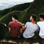 four men with lighter skin tones sitting on the ground, and looking over lush, green hills and laughing